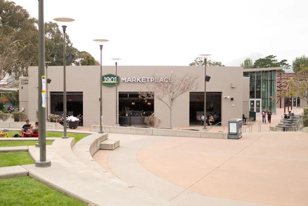 Students gather outside the 1901 Marketplace building at Cal Poly, featuring outdoor seating, a courtyard with pathways, and the marketplace’s modern exterior.