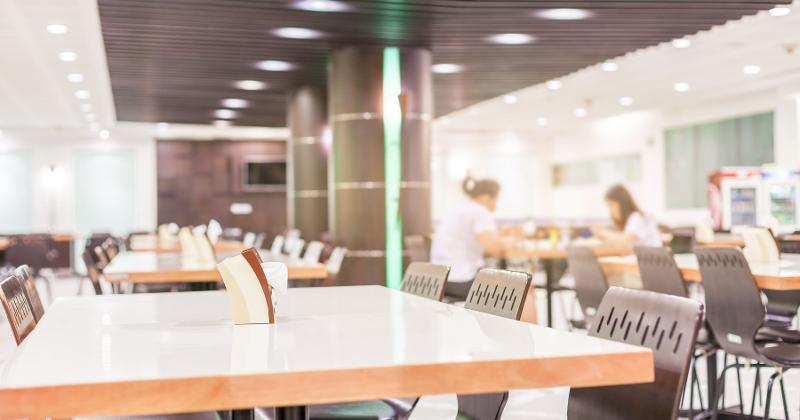 Bright, modern dining hall interior with rows of empty tables and chairs, softly lit and featuring two people seated in the background near a food service area.