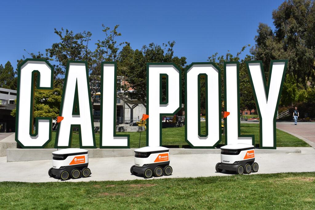 Three Grubhub Starship delivery robots with orange flags drive past the large green-and-white “CAL POLY” sign on campus under a clear blue sky.