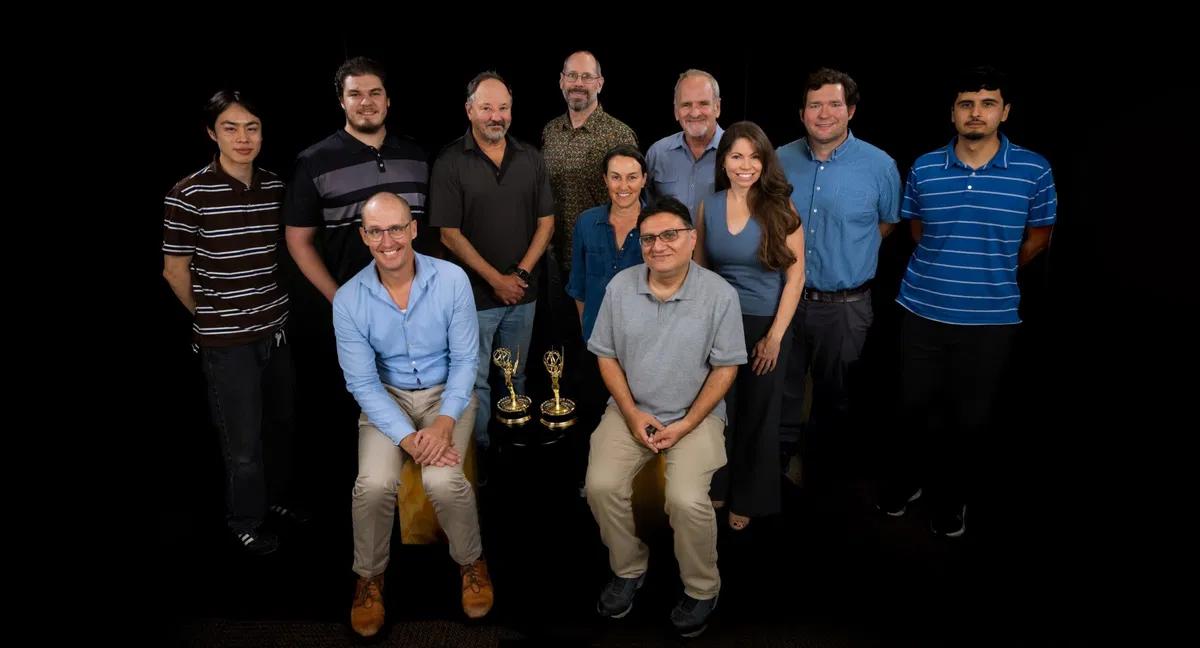 Group photo of a team posing together in front of a black background with two Emmy Awards displayed on a table in front of them.