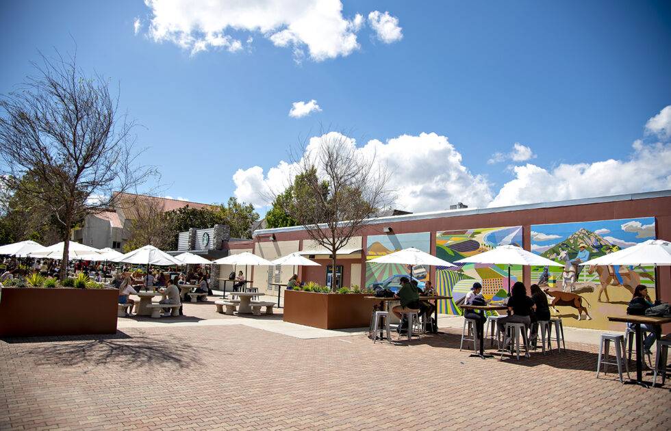 Students sitting under umbrellas at outdoor tables near a colorful mural on a sunny campus patio.