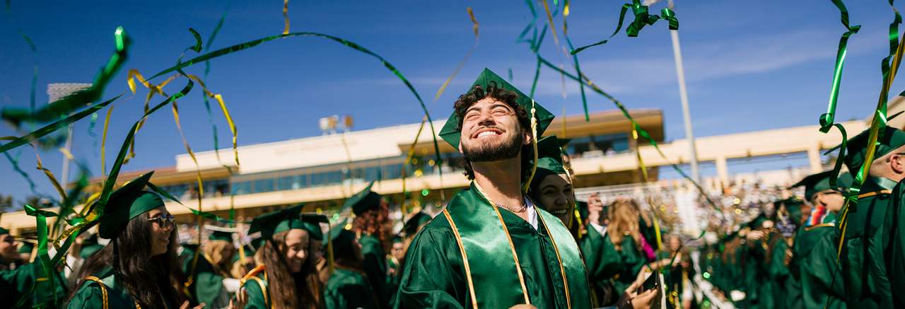 Graduates in green caps and gowns celebrate at an outdoor commencement ceremony with gold and green streamers flying through the air under a clear blue sky.