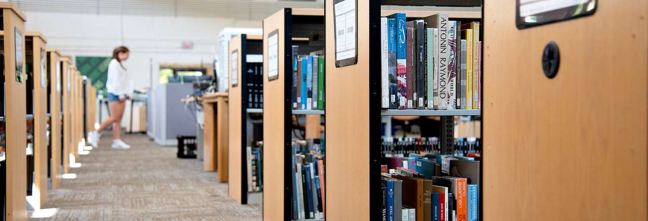 Bookshelves filled with books in a library aisle, with a student standing in the background near a counter or workstation.