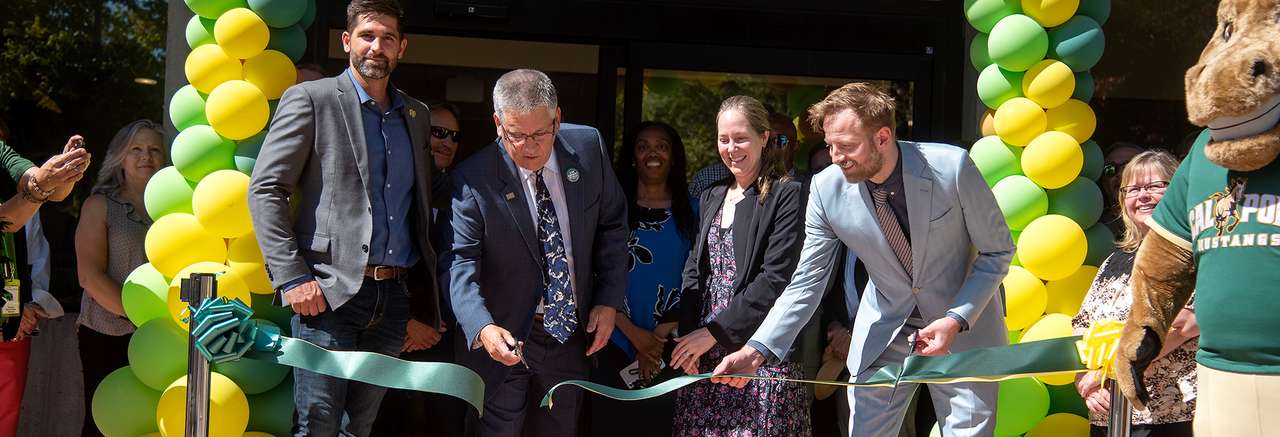 Officials and guests participate in a ribbon-cutting ceremony for the Mustang Shop, surrounded by green and yellow balloons, with the Cal Poly Mustang mascot standing nearby.