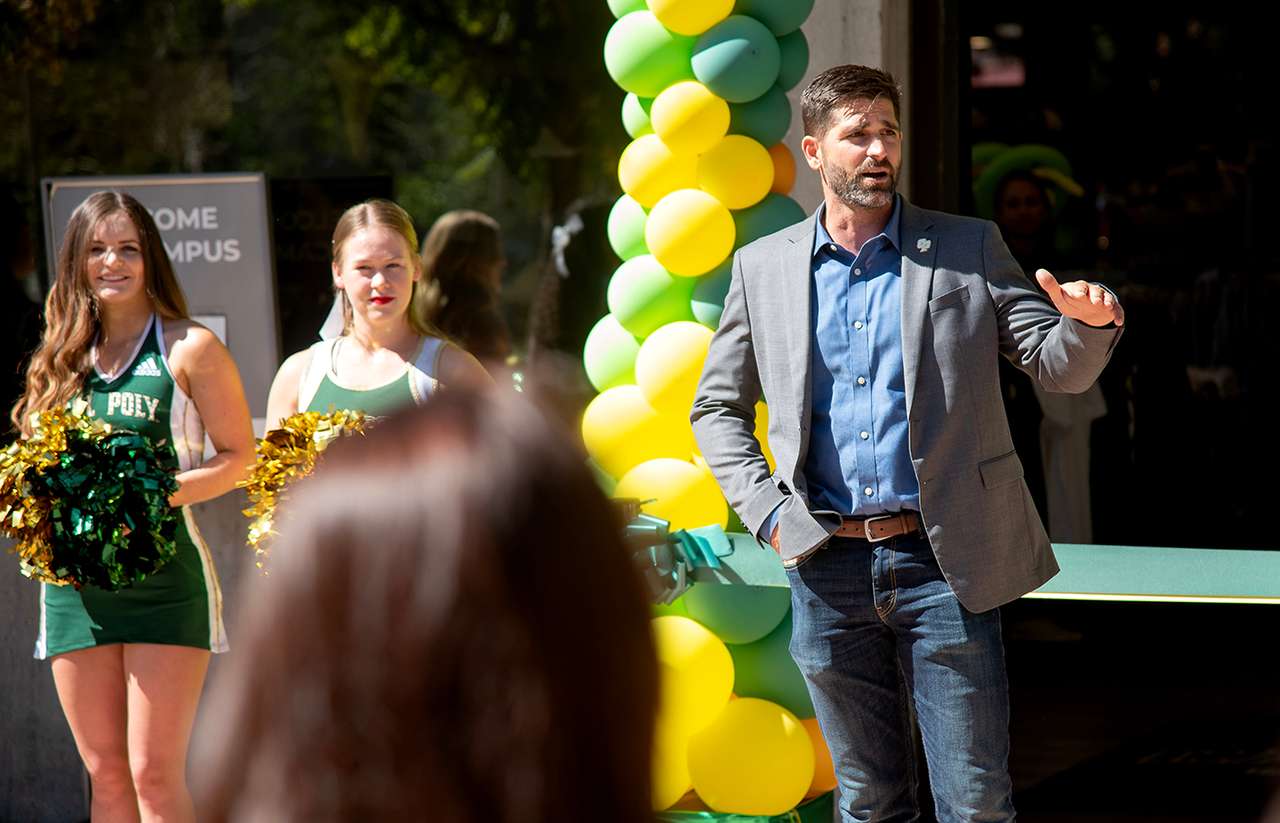A man in a blazer speaks at a ribbon-cutting ceremony, flanked by Cal Poly cheerleaders and a green and yellow balloon arch in the background.