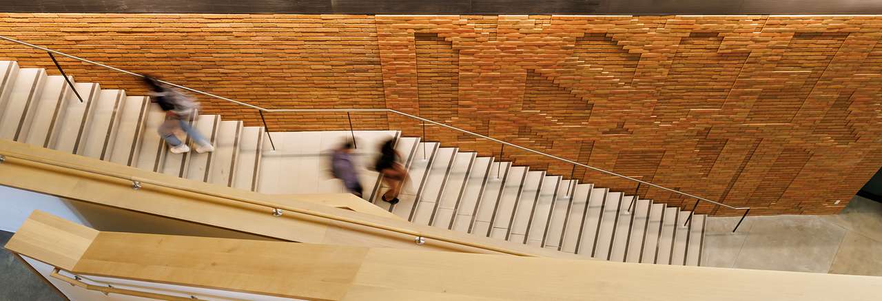 Blurred students walk down a modern staircase inside a building with a prominent red brick wall featuring abstract geometric patterns.