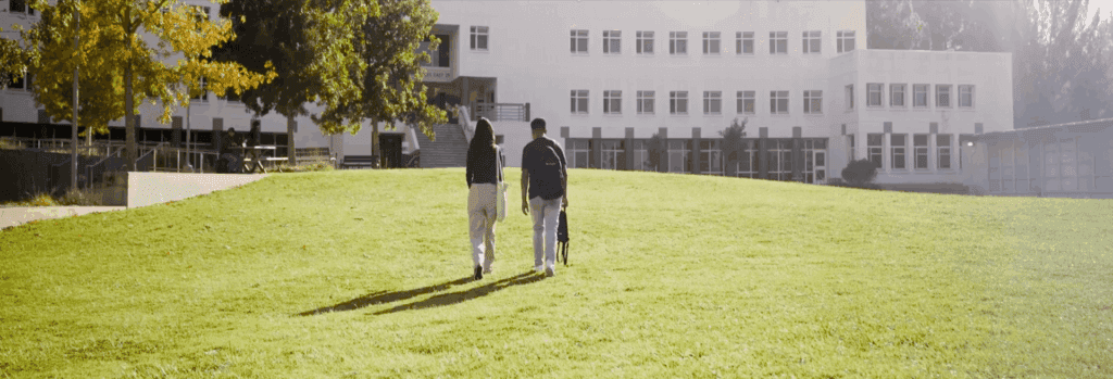 Two students walk side-by-side across a sunlit grassy lawn toward a modern academic building with large windows and trees showing fall colors.
