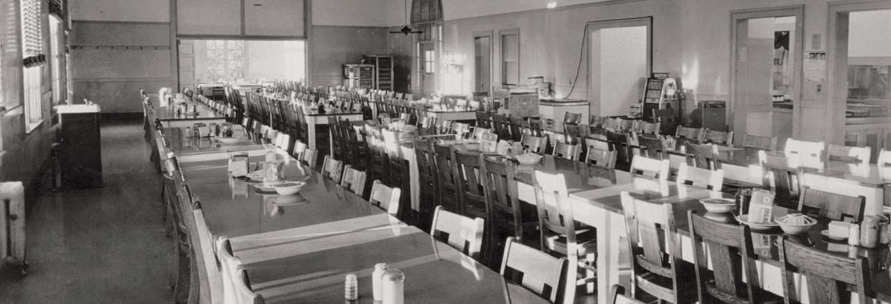 Black and white historical photo of a large dining hall with rows of wooden tables and chairs set for a meal, likely from a mid-20th century institutional setting.